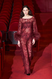 Model on a runway wearing a burgundy lace jumpsuit with oversized feathered cuffs inside a red-seated theater venue
