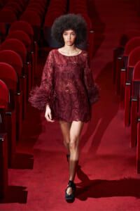 Female model walks down a red theater aisle in a burgundy lace dress with feathered cuffs.