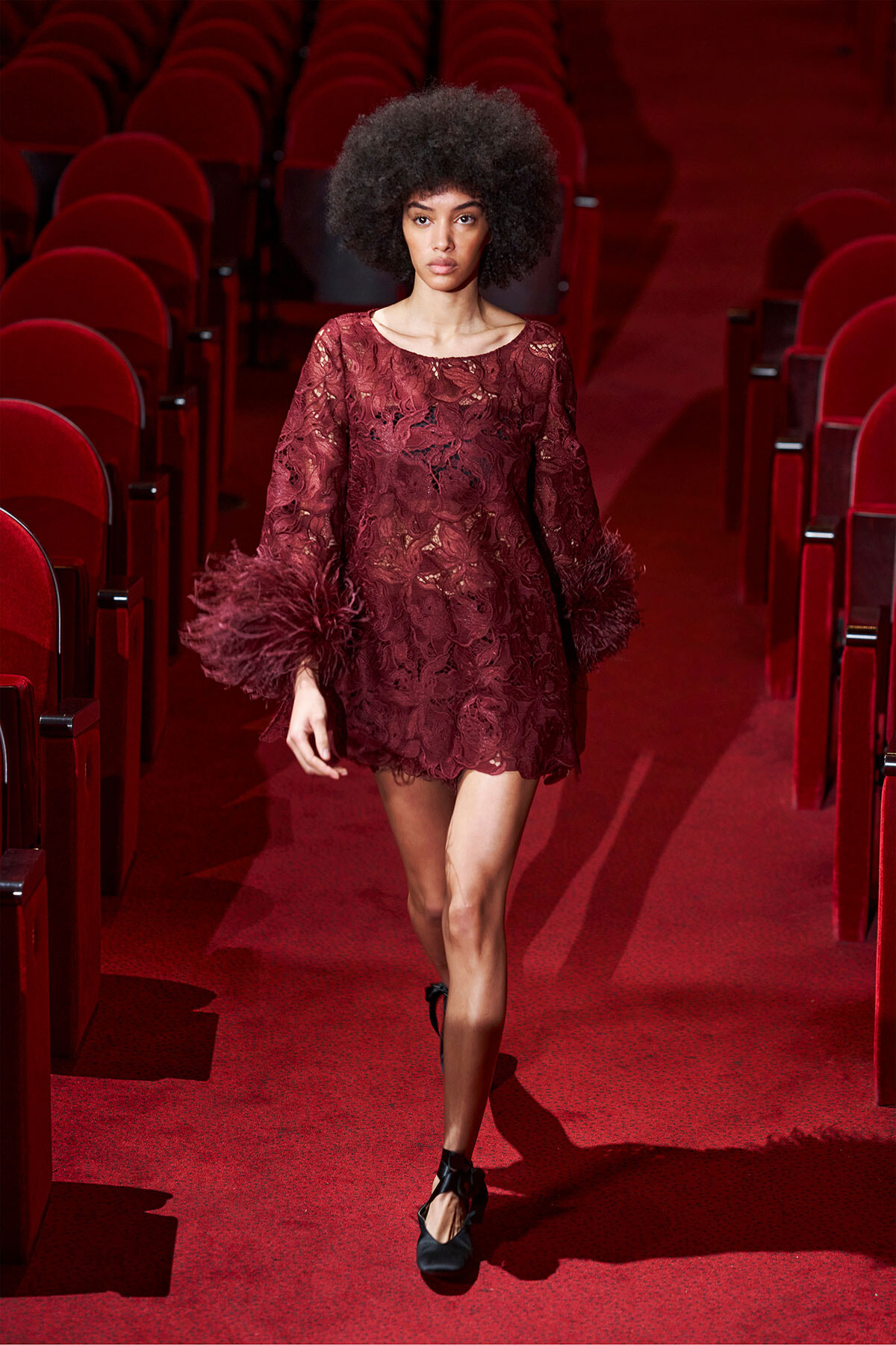 Female model walks down a red theater aisle in a burgundy lace dress with feathered cuffs.