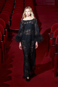 Model walking down a red-carpeted theater aisle in a long black lace dress with feathered cuffs.