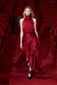 Model walking down a red-theater aisle in a burgundy halter-neck gown with layered ruffles and black ribbon-tied flats.