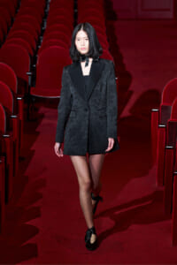 Model walking down a red theater aisle in a black textured blazer dress with a satin lapel and bow choker.