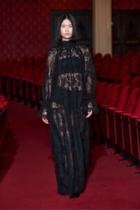 Model walking down a red theater aisle in a full-length black lace gown with a high neck and sheer sleeves.