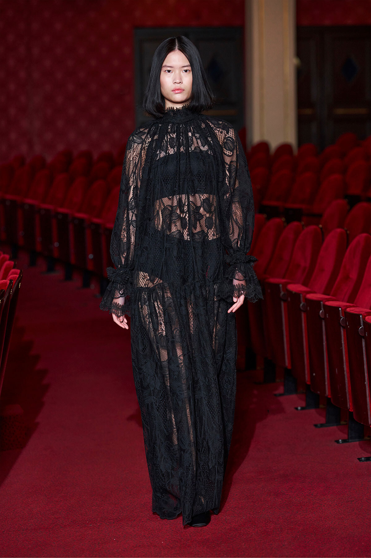 Model walking down a red theater aisle in a full-length black lace gown with a high neck and sheer sleeves.
