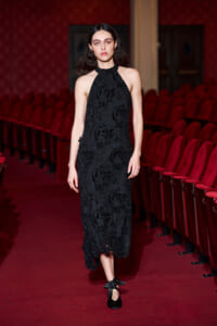 Model wearing a black lace halter midi dress walking along a theater aisle with red upholstered seats in the background.