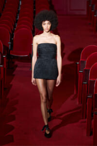 Model walking down a red theater aisle in a strapless black mini dress with black heels with ribbons at the ankles.
