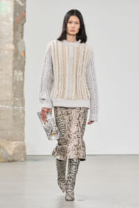 Model wearing a cream chunky-knit sweater and snake-print midi skirt, carrying a snakeskin clutch and matching boots on a runway.