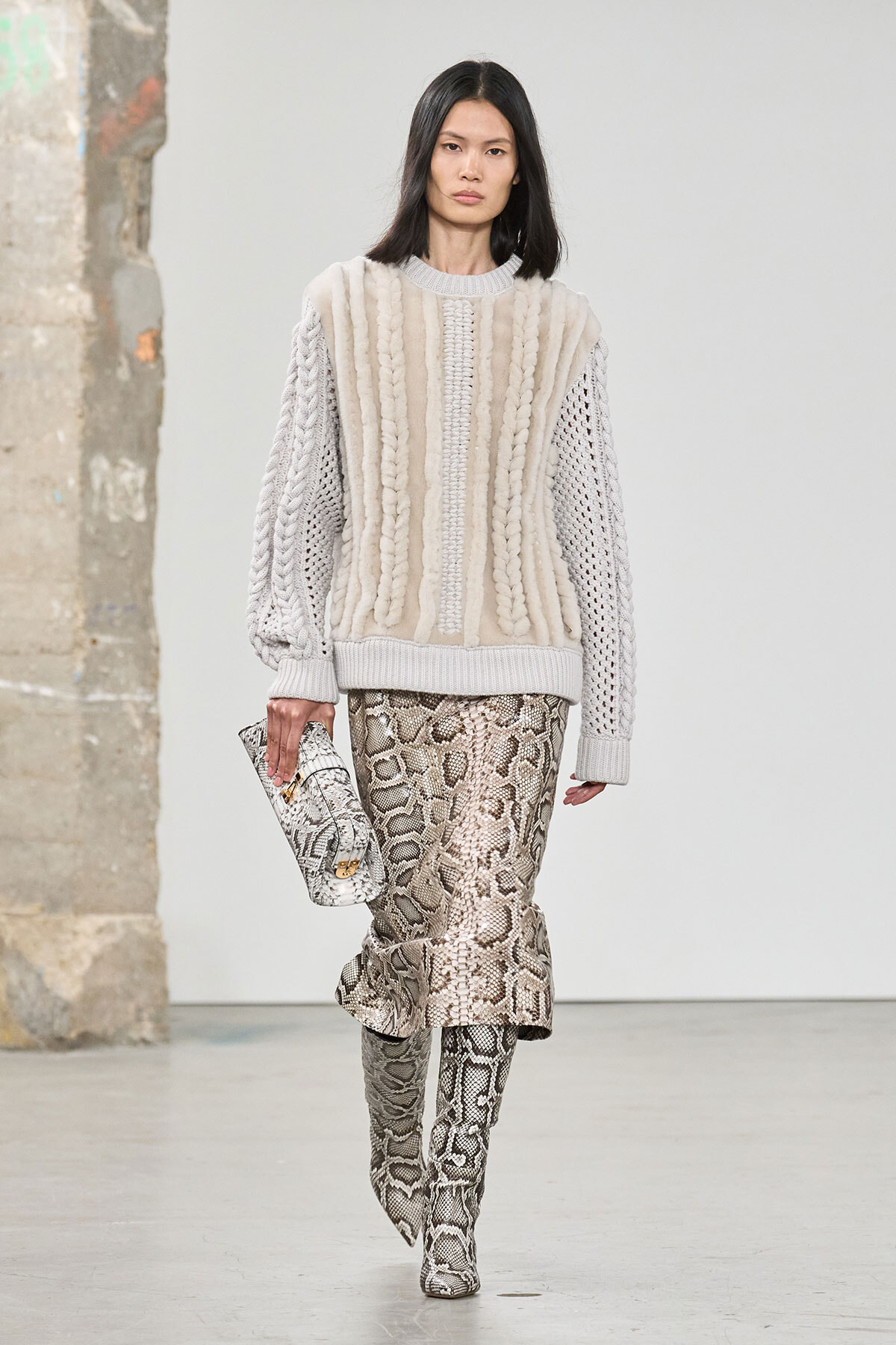 Model wearing a cream chunky-knit sweater and snake-print midi skirt, carrying a snakeskin clutch and matching boots on a runway.