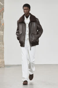 Male model walking the runway in a dark brown shearling-collar leather bomber jacket, white layers, and brown shoes.