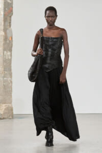 Runway model walking in a black textured strapless top, flowing black skirt, black boots, and a large woven shoulder bag.