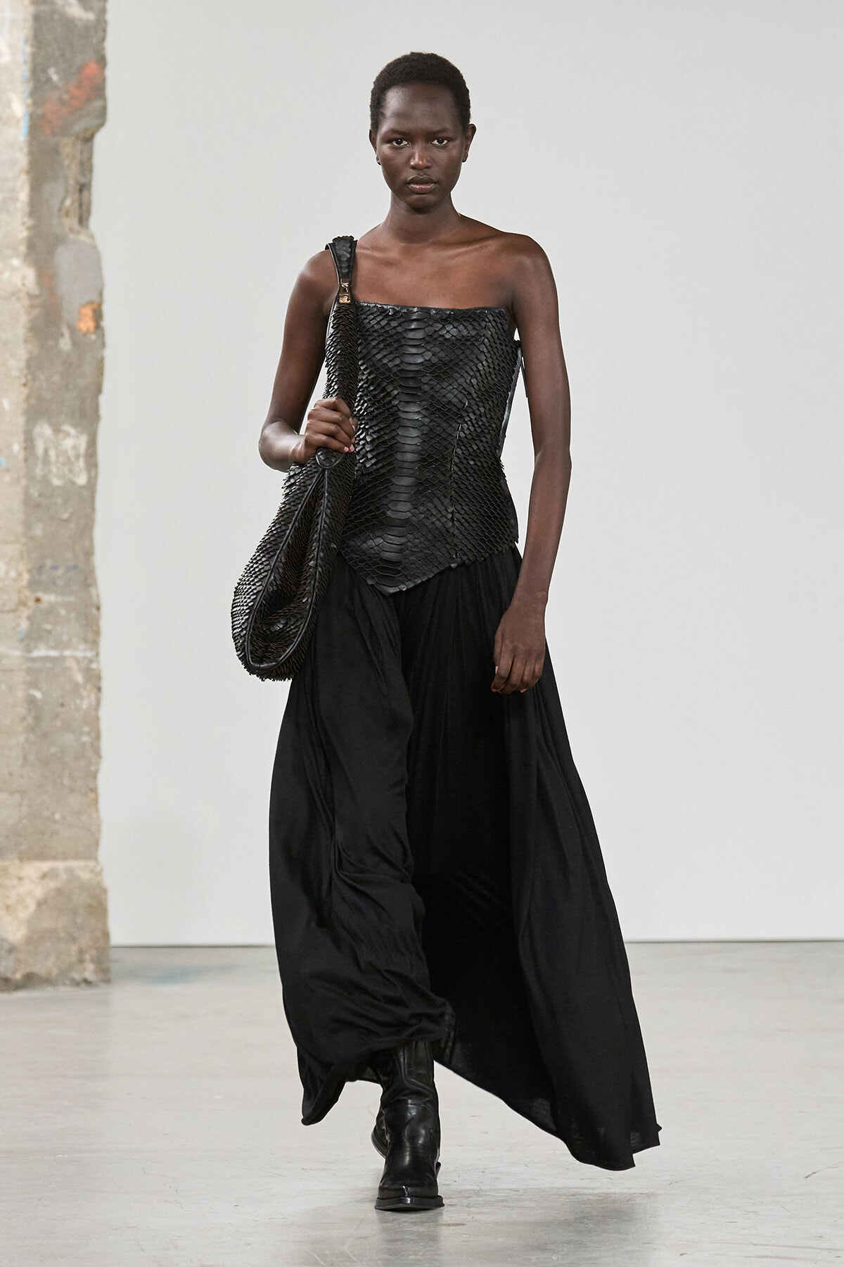 Runway model walking in a black textured strapless top, flowing black skirt, black boots, and a large woven shoulder bag.