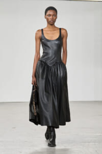 Model walking in a black leather sleeveless gown with a gathered midi skirt, holding a small chain-strap bag and wearing ankle boots.