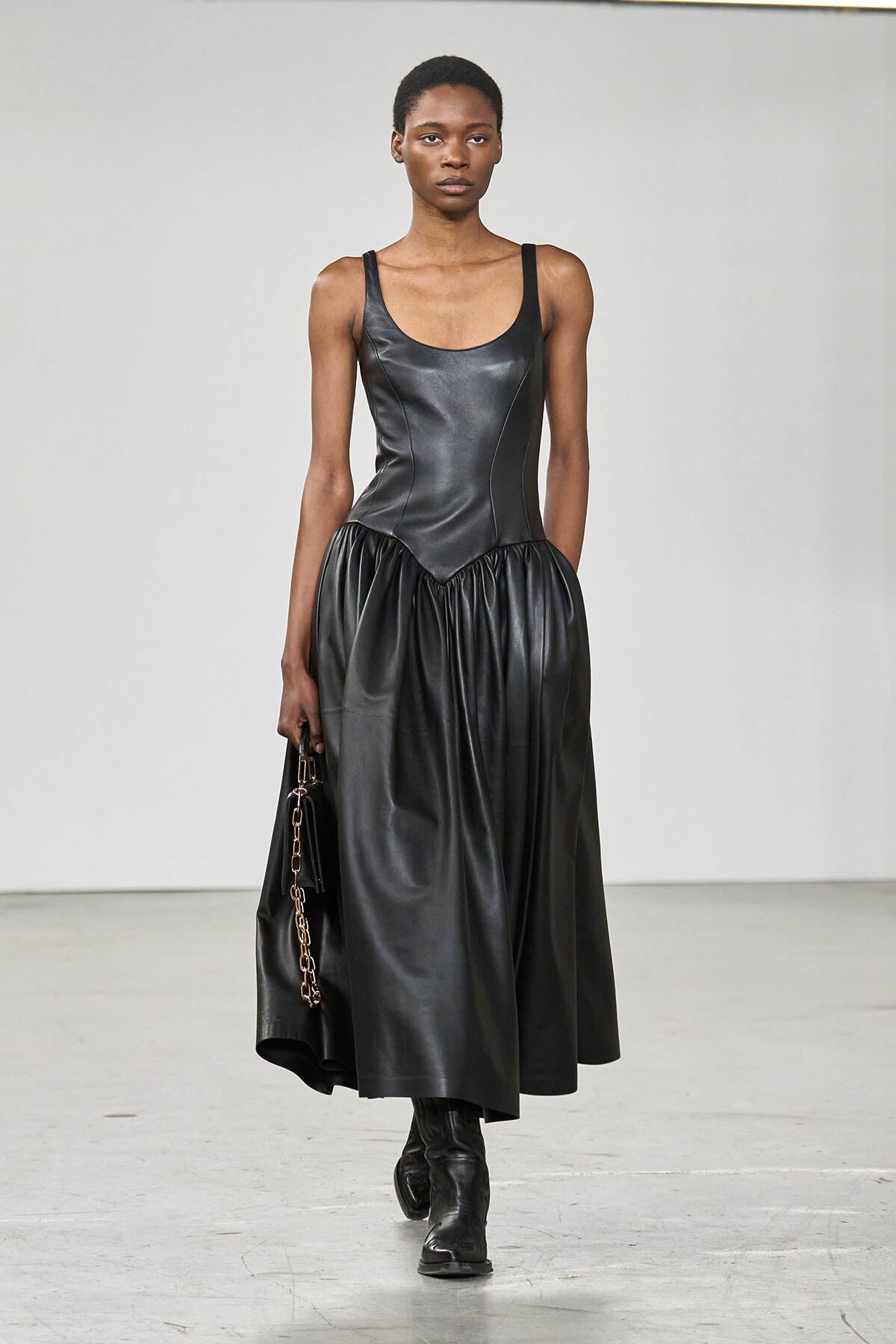 Model walking in a black leather sleeveless gown with a gathered midi skirt, holding a small chain-strap bag and wearing ankle boots.