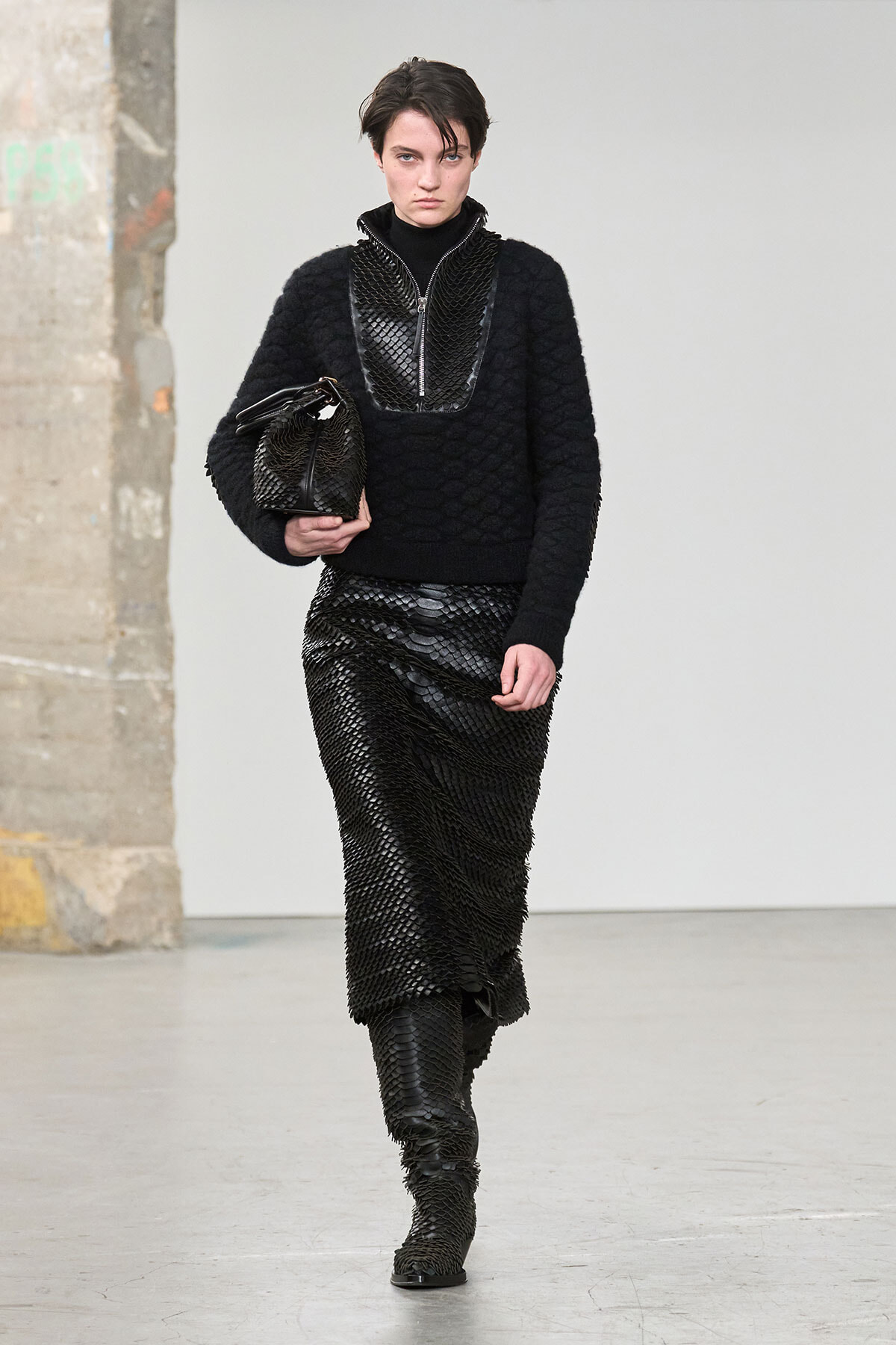 Female model walking in a black textured knit sweater and scale-pattern skirt, carrying a matching black handbag.