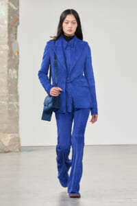 Model walking in a cobalt-blue suit with blazer and matching pants, carrying a blue crossbody bag.