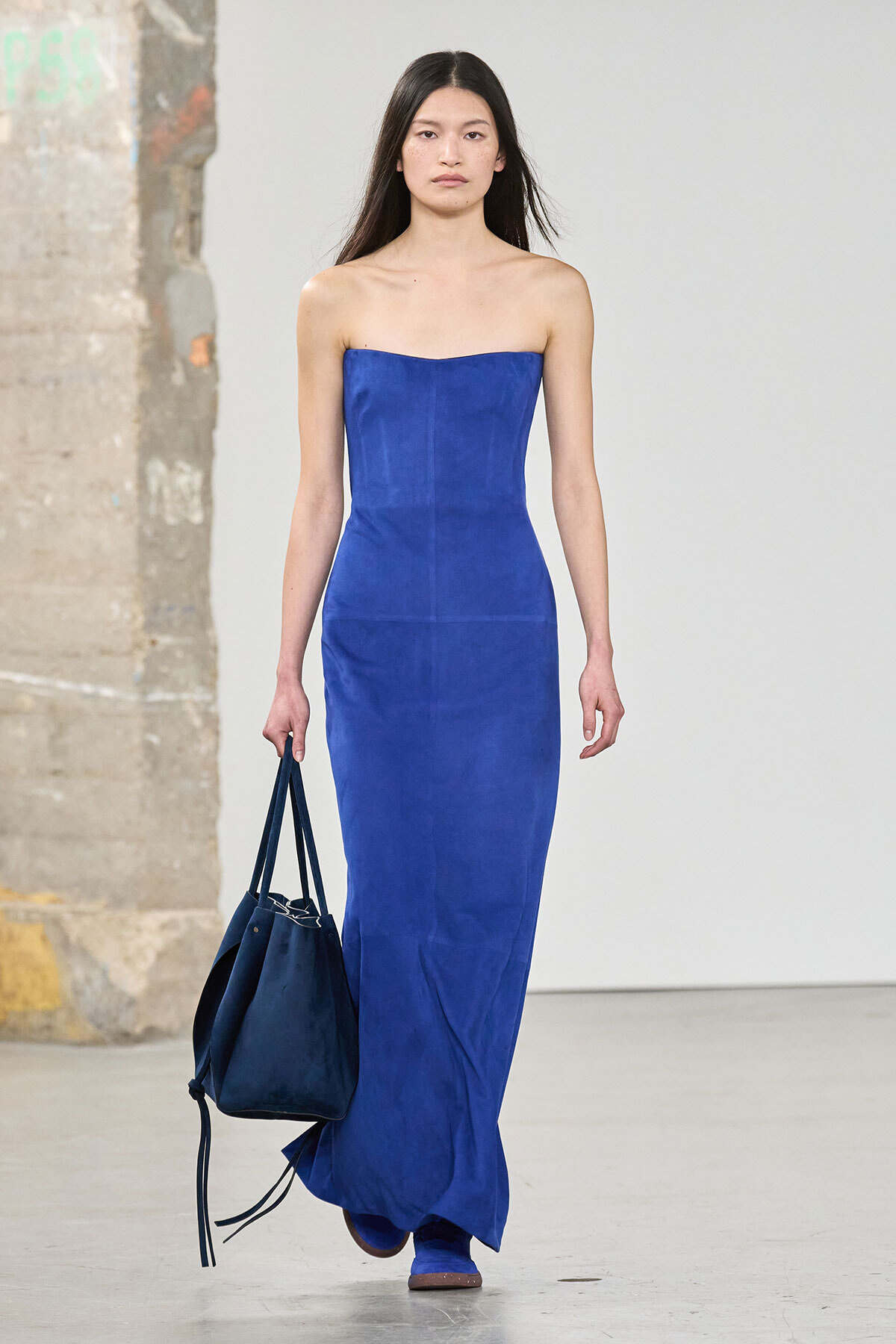 Model on a runway wearing a cobalt blue strapless maxi dress, carrying a navy tote bag.