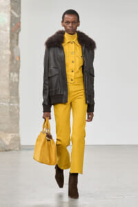 Model walking on a runway in a mustard-yellow jumpsuit with a black fur-trim jacket and brown ankle boots, carrying a matching yellow bag