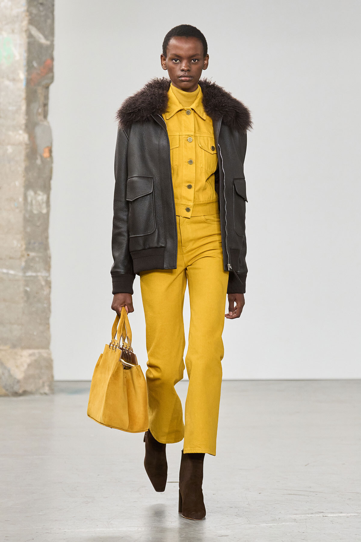 Model walking on a runway in a mustard-yellow jumpsuit with a black fur-trim jacket and brown ankle boots, carrying a matching yellow bag