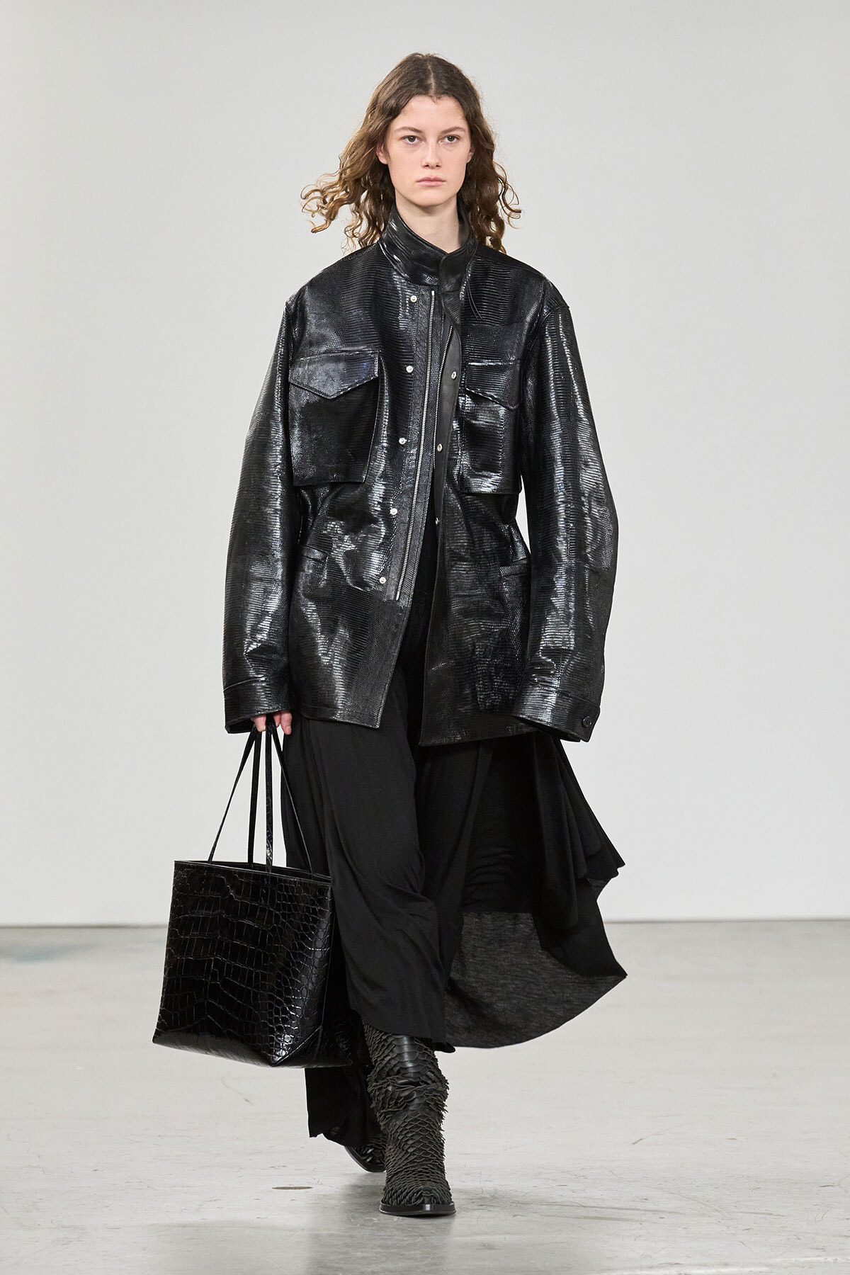 Model on a runway wearing an oversized black metallic jacket with large pockets, a long black skirt, textured black boots, and carrying a black crocodile-textured tote bag.