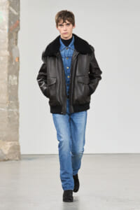 Male model walking a runway in a black leather jacket with a shearling collar over a blue denim shirt and jeans, hands in pockets.