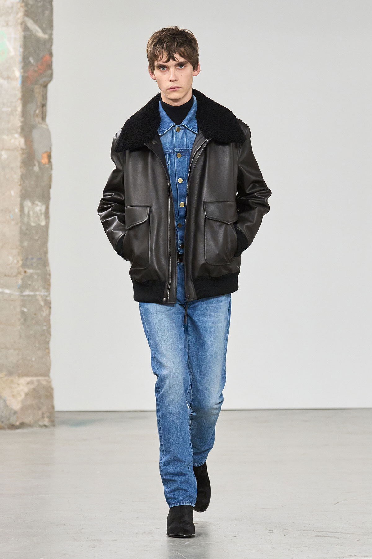 Male model walking a runway in a black leather jacket with a shearling collar over a blue denim shirt and jeans, hands in pockets.