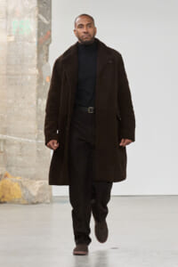 Male model wearing a long brown shearling coat over a black turtleneck and trousers, walking on a fashion runway.