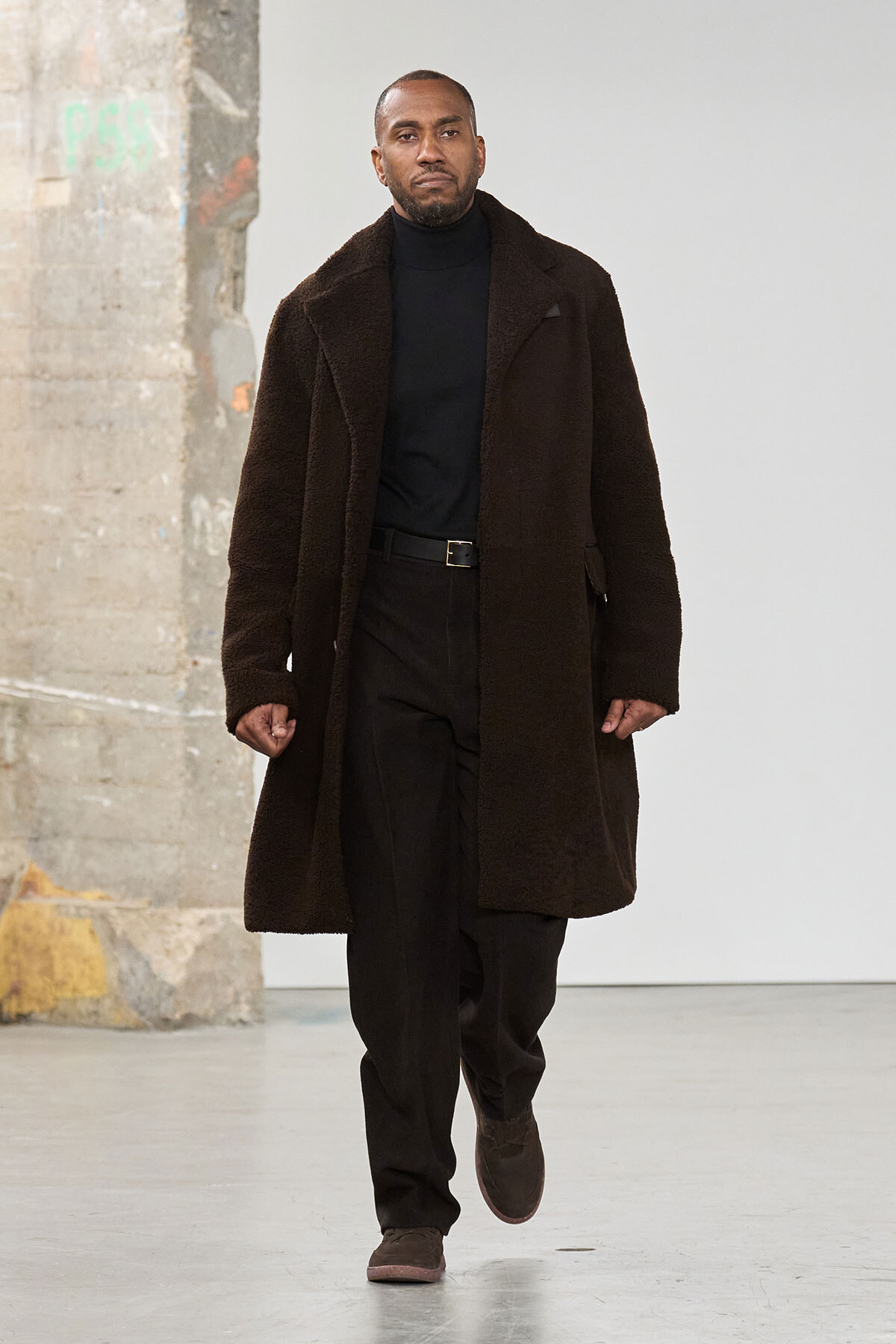 Male model wearing a long brown shearling coat over a black turtleneck and trousers, walking on a fashion runway.