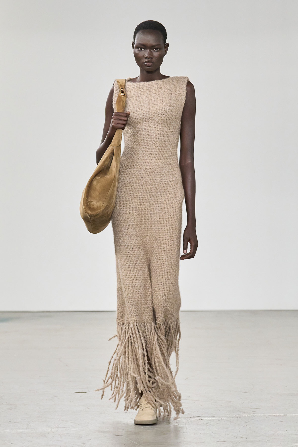 Model walks the runway in a sleeveless beige textured knit dress with a long fringed hem, carrying a large tan shoulder bag.