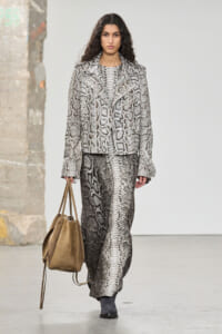 Model walks the runway in a gray snakeskin-print blazer and matching long skirt, carrying a tan suede tote.