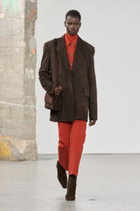 Model walking the runway in an oversized brown blazer with burnt orange turtleneck and matching orange-red pants, carrying a small brown crossbody bag.