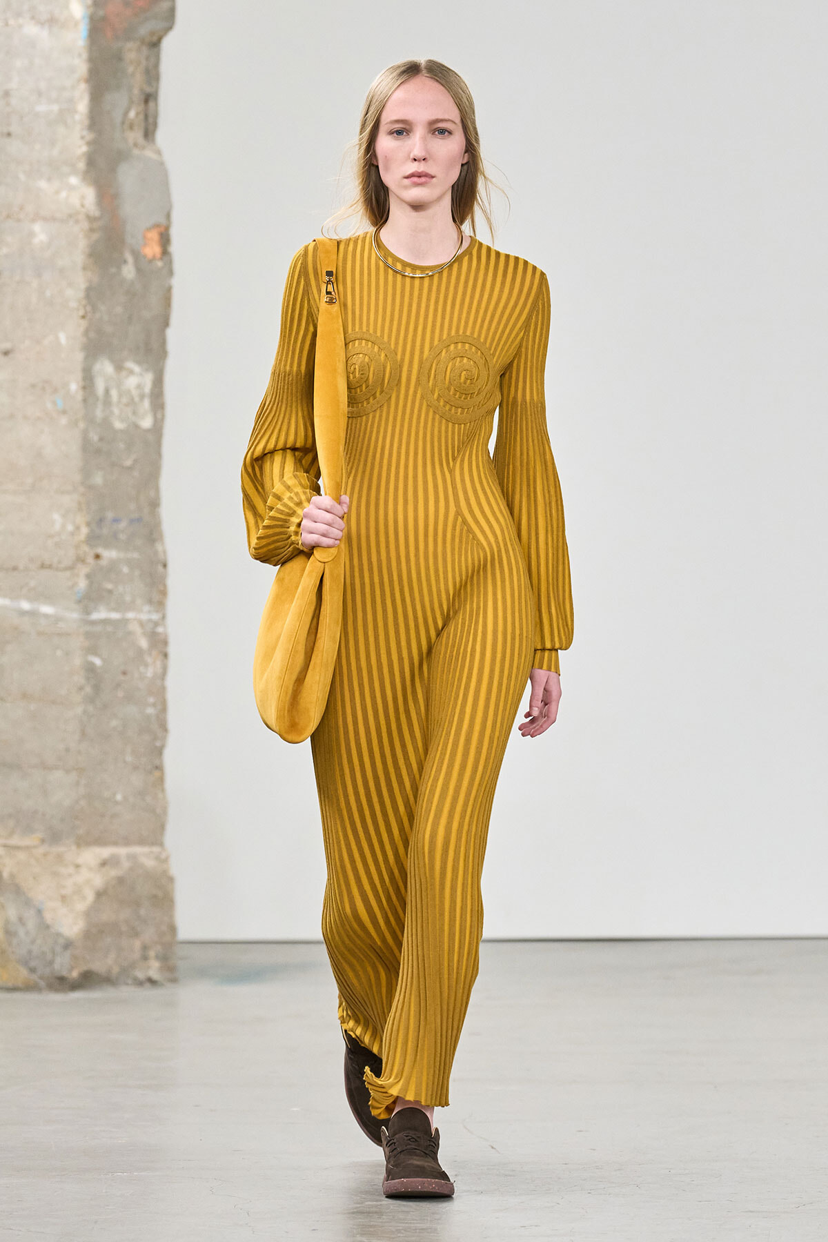 Model walking runway in mustard-yellow striped jumpsuit with matching shoulder bag and brown shoes.
