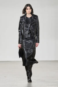 Model walking a runway in a shiny black patent leather jacket and matching long skirt with knee-high boots, holding a small dark clutch.