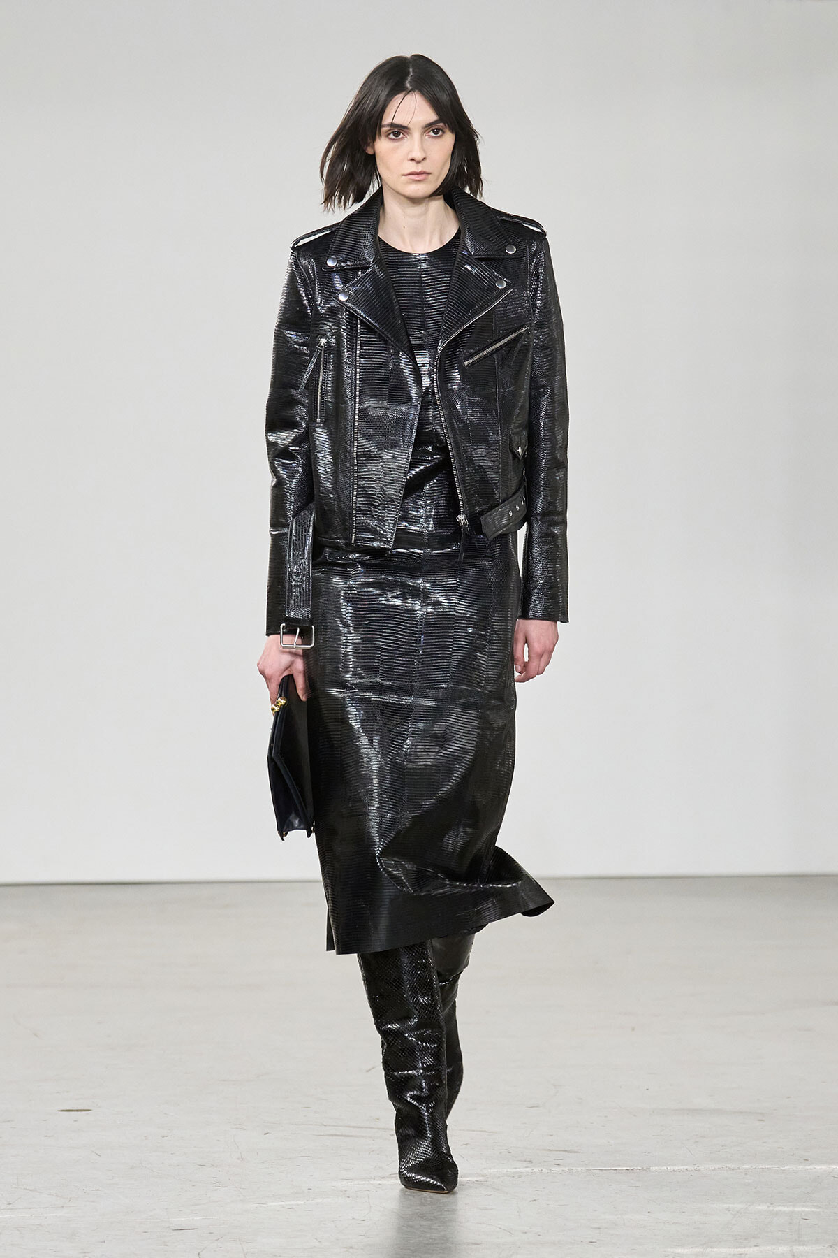 Model walking a runway in a shiny black patent leather jacket and matching long skirt with knee-high boots, holding a small dark clutch.