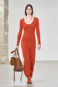 Model walking runway in an orange knit jumpsuit with matching orange shoes, carrying two brown bags and wearing a spiky necklace.