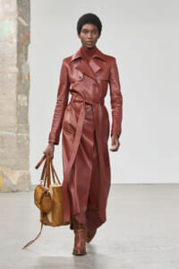 Model wearing a burgundy leather trench coat with a belt, paired with matching boots and a tan shearling handbag.