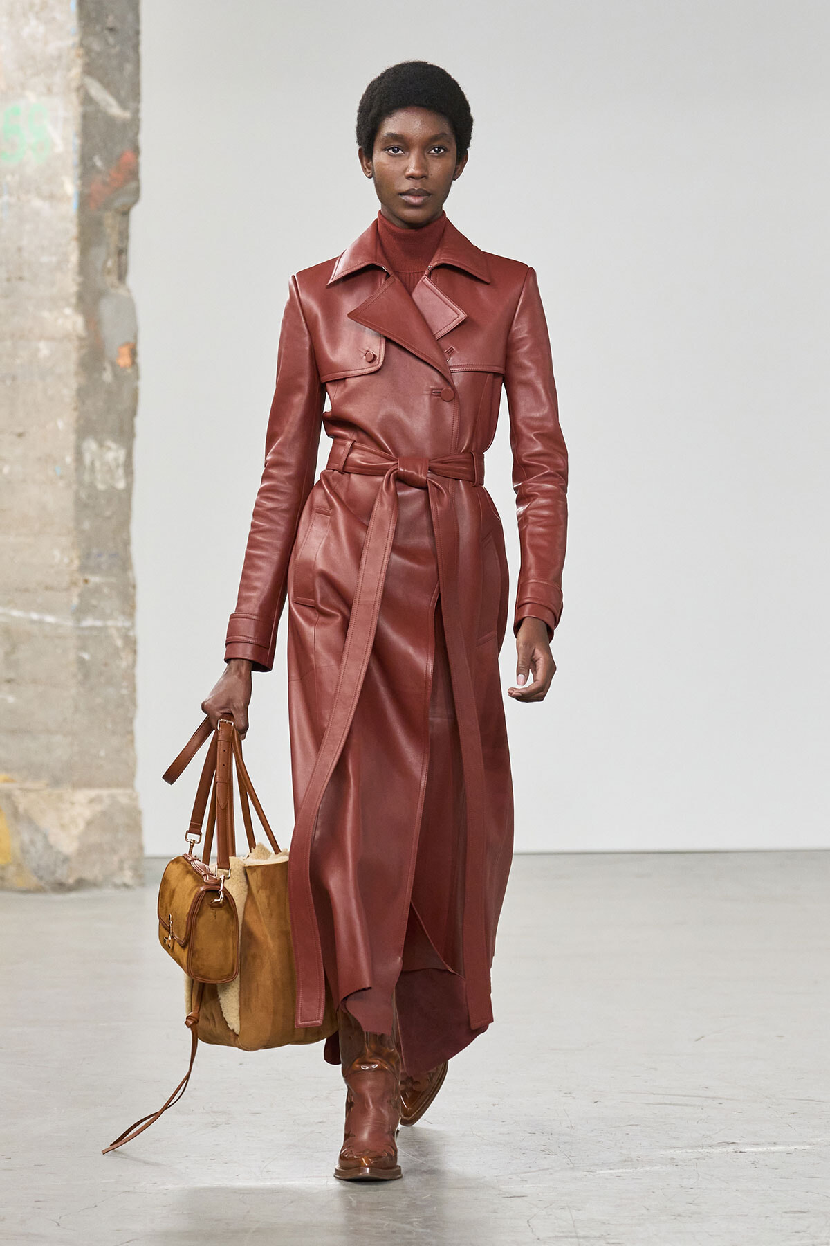 Model wearing a burgundy leather trench coat with a belt, paired with matching boots and a tan shearling handbag.