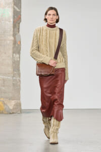 Model walking a fashion runway in a chunky beige knit sweater, burgundy leather skirt, and cream knitted boots, carrying a brown crossbody bag with a chain strap.