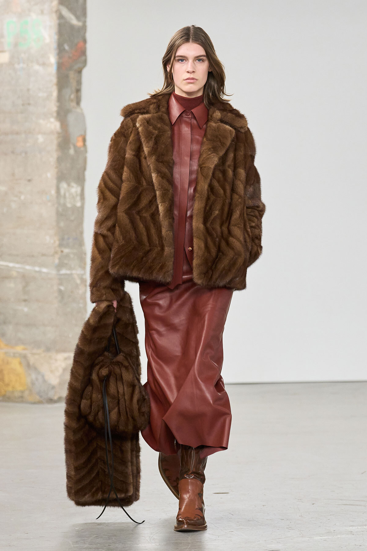 Female model on a runway wearing a brown fur coat over a burgundy leather outfit, knee-high boots, and carrying a matching fur bag.