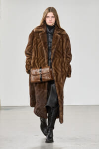 Model walking a runway in a long brown fur coat over black leather, paired with a brown snakeskin handbag with a black strap.