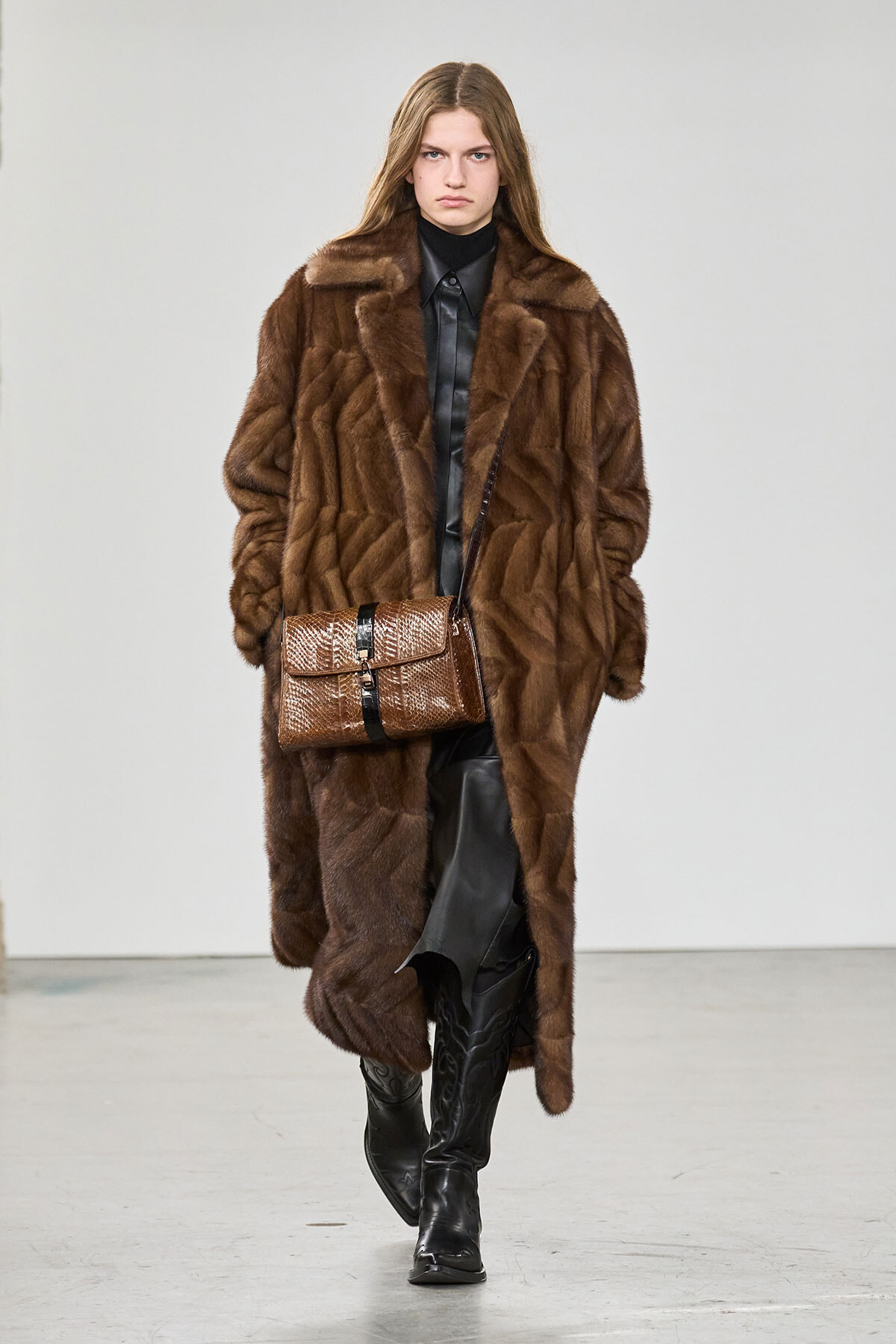 Model walking a runway in a long brown fur coat over black leather, paired with a brown snakeskin handbag with a black strap.