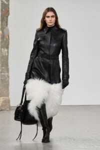 Model walking in a black leather belted coat with pockets, paired with white fur leg coverings and knee-high boots, carrying a black handbag.