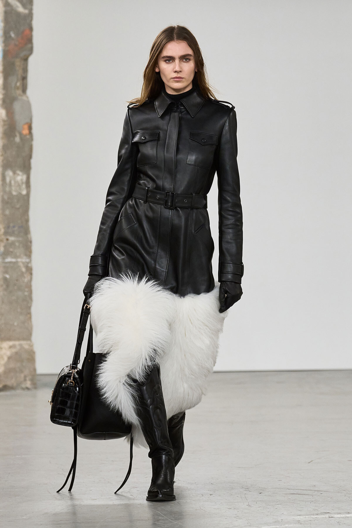 Model walking in a black leather belted coat with pockets, paired with white fur leg coverings and knee-high boots, carrying a black handbag.