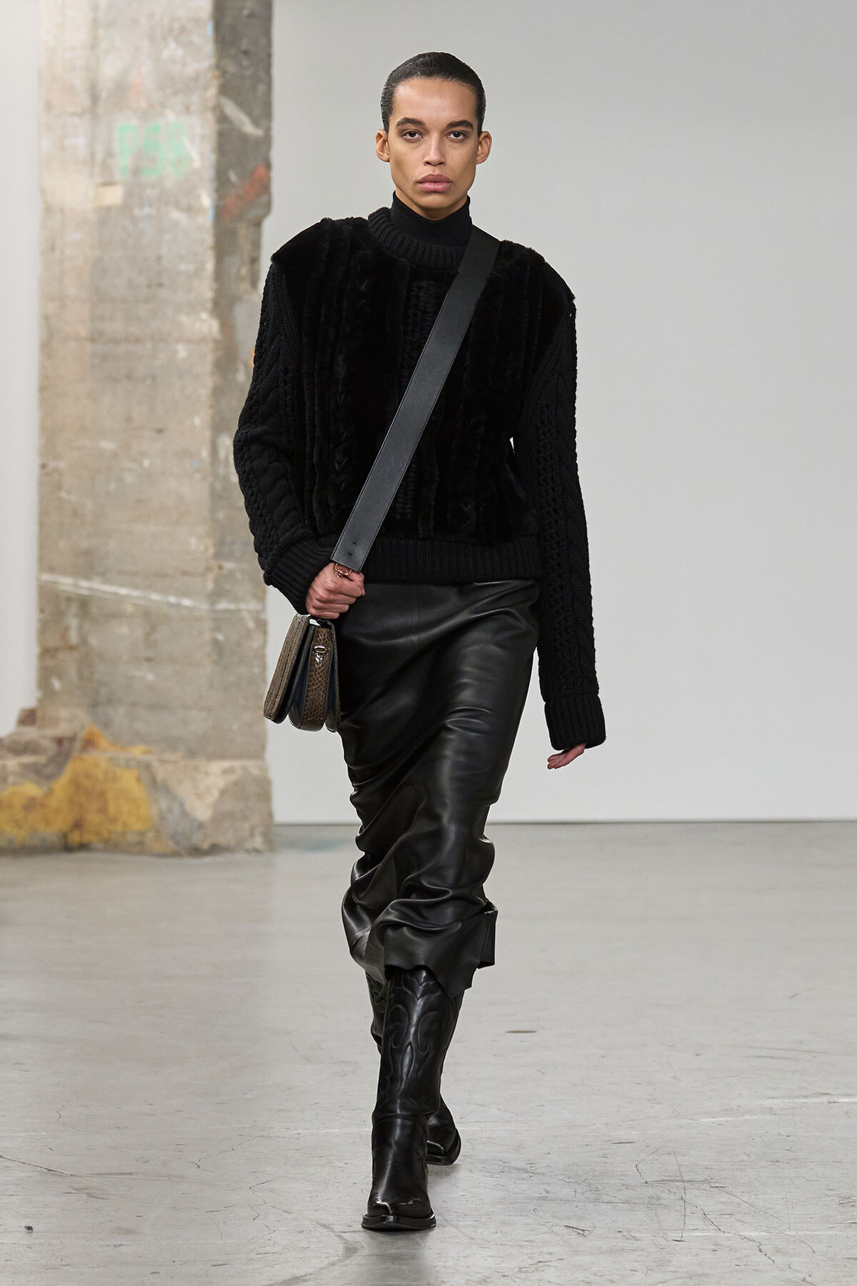 Model walking in an all-black outfit: textured knit sweater, glossy leather midi skirt, knee-high boots, and a crossbody bag with a wide strap.