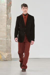 Male model walking a runway in a dark corduroy blazer over a rust-red shirt and pants, brown shoes.