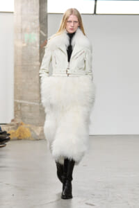 Model walking a runway in an ivory fur coat with a matching fluffy skirt, belt, and black knee-high boots in an industrial venue.