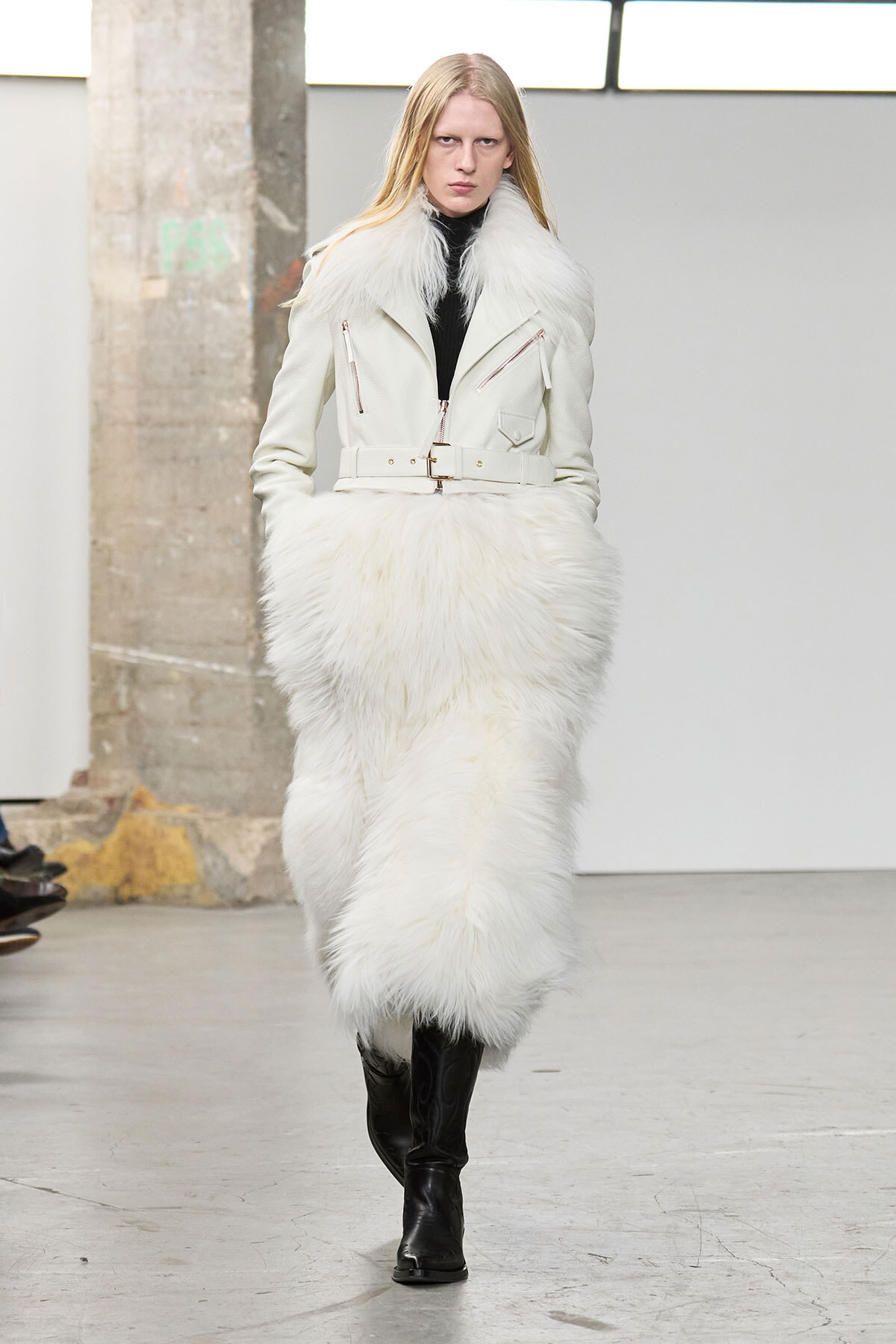Model walking a runway in an ivory fur coat with a matching fluffy skirt, belt, and black knee-high boots in an industrial venue.