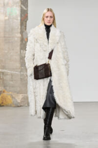 Model on a fashion runway wearing a long white shaggy fur coat over black, with knee-high boots and a dark brown textured handbag.