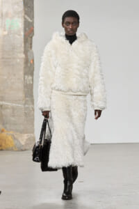 Model walking a runway in a white fluffy zippered coat, black turtleneck, knee-high boots, and a black handbag.