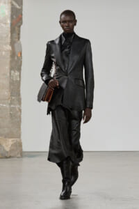 Model wearing a head-to-toe black leather suit with an oversized blazer, black shirt, and knee-high boots, carrying a small brown chain-strap bag regretfully.