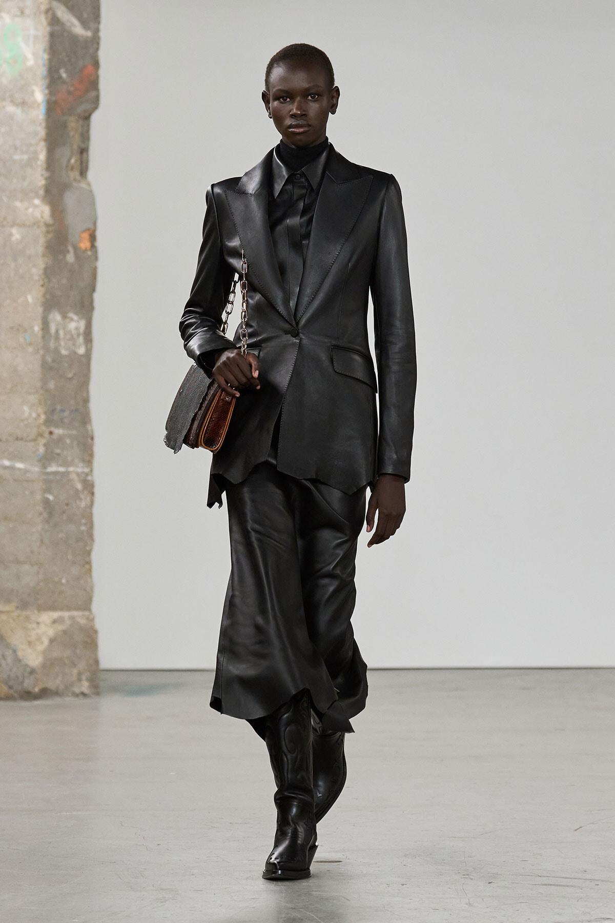Model wearing a head-to-toe black leather suit with an oversized blazer, black shirt, and knee-high boots, carrying a small brown chain-strap bag regretfully.